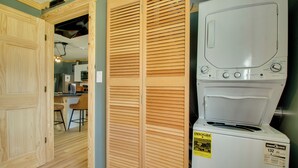 Une buanderie équipée d’un lave-linge à chargement frontal, de portes de placard en bois à persiennes et d’un coin cuisine visible à travers une porte ouverte.
