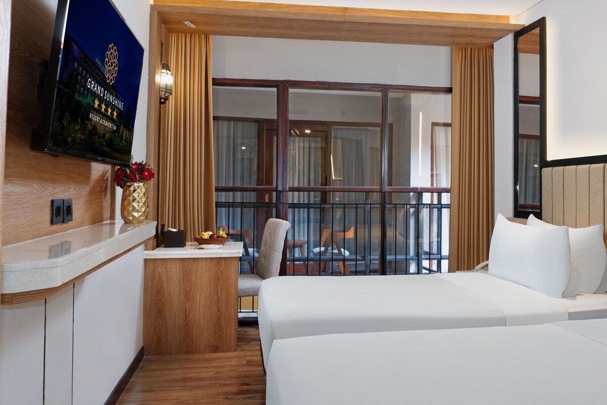 A hotel room with a bed, a desk with a TV, a window with curtains, and a view of the outside.