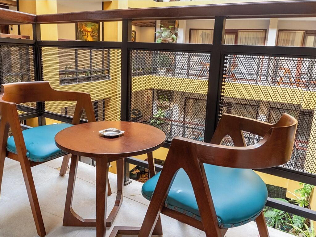 Un balcón con dos sillas de madera y una mesa, un separador de malla y vista a la calle.