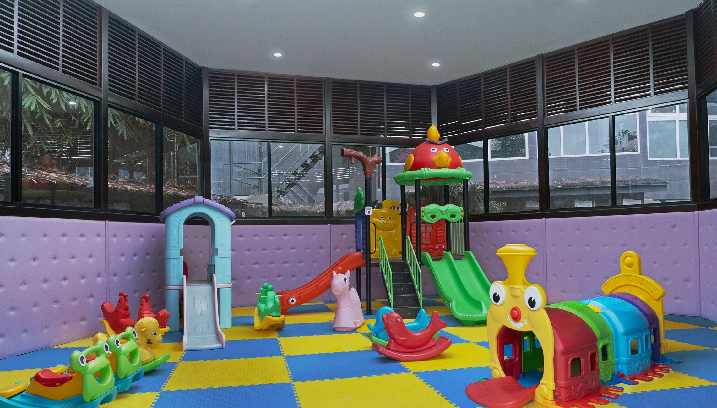A children's play area with various colorful play structures, including slides and climbing frames, set against a purple wall with large windows.