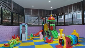 A children's play area with various colorful play structures, including slides and climbing frames, set against a purple wall with large windows.