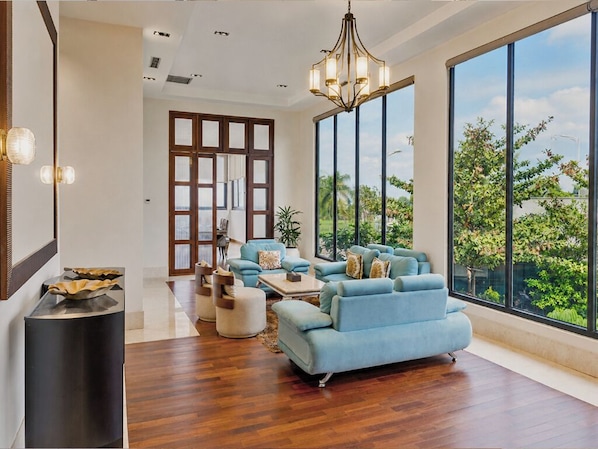 A spacious living room with a large window, a blue sofa set, and a modern chandelier.