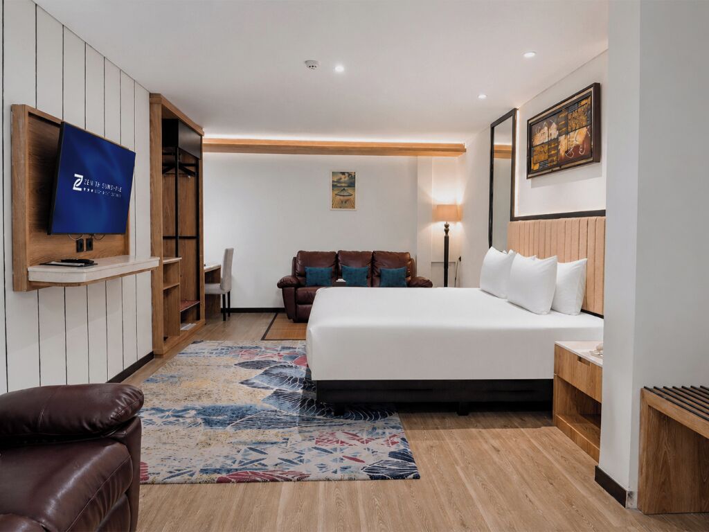 Quarto de hotel com cama, televisão na parede, sofá, luminária e um quadro na parede.