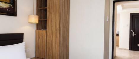 A hotel room with a bed, a wooden wardrobe, a wall-mounted lamp, and a door leading to another room.