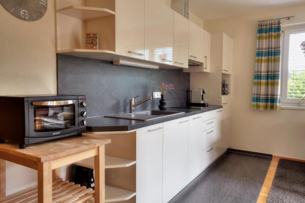 A modern kitchen with white cabinets, a black countertop, a built-in oven, and a window with a striped curtain.