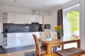A modern kitchen with white cabinets, a dining area with a wooden table and wicker chairs, and a view of greenery outside. - Hölderle Damüls (Damüls)