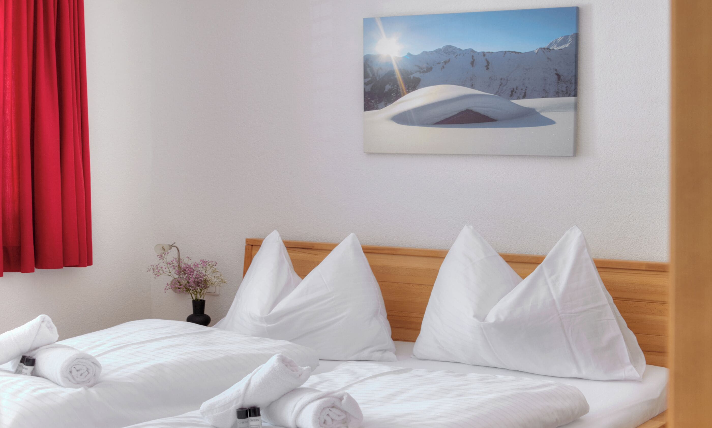 Habitación de hotel con cama, almohadas, un cuadro de un paisaje nevado de montaña, un florero con flores y una cortina roja.
