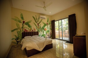 Quarto com cama, ventilador de teto, ar condicionado, porta de correr de vidro para uma varanda e uma planta.