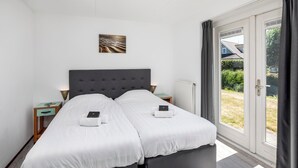 En dobbeltseng med gråt polstret sengegavl, to sengeborde med lamper, en radiator og udsigt til det fri gennem en skydedør.