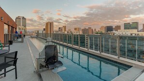 Une piscine sur le toit, entourée d’une paroi en verre, avec un barbecue, une table et des chaises.
