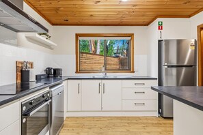 A modern kitchen with white cabinets, stainless steel appliances, a wooden ceiling, and a window with a view of trees.