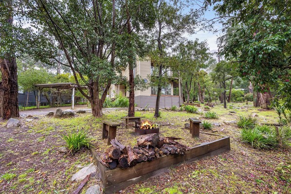 A wooden deck with a fire pit, surrounded by trees and a building in the background.