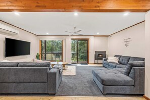 A modern living room with a flat-screen TV, grey sofas, a fireplace, and a large window with a view of greenery. - Gariwerd Family Lodge (Halls Gap)