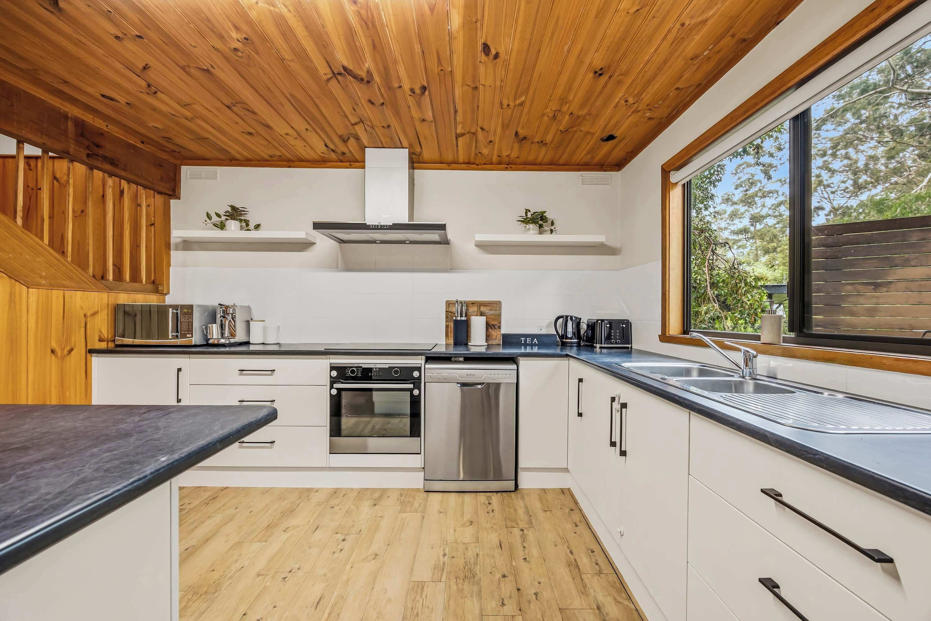 A modern kitchen with wooden ceiling beams, white cabinetry, a built-in oven, and a large window offering a view of the outdoors.