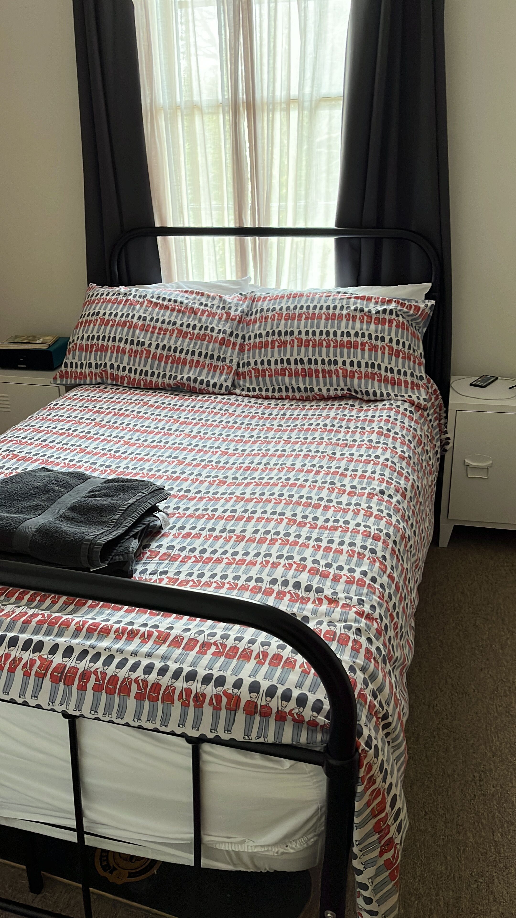 Una cama con colcha estampada, estructura metálica negra, mesita de noche con teléfono y ventana con cortinas translúcidas.