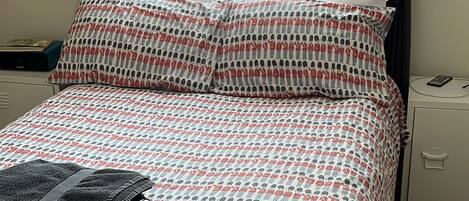 Una cama con colcha estampada, estructura metálica negra, mesita de noche con teléfono y ventana con cortinas translúcidas.