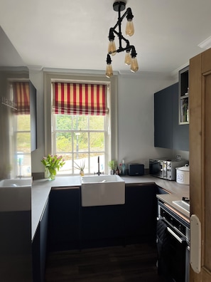 A modern kitchen with a built-in sink, a window with a striped valance, and a pendant light fixture. - North Mill Cottage | Charming Cottage at Cowdray Park (Midhurst)