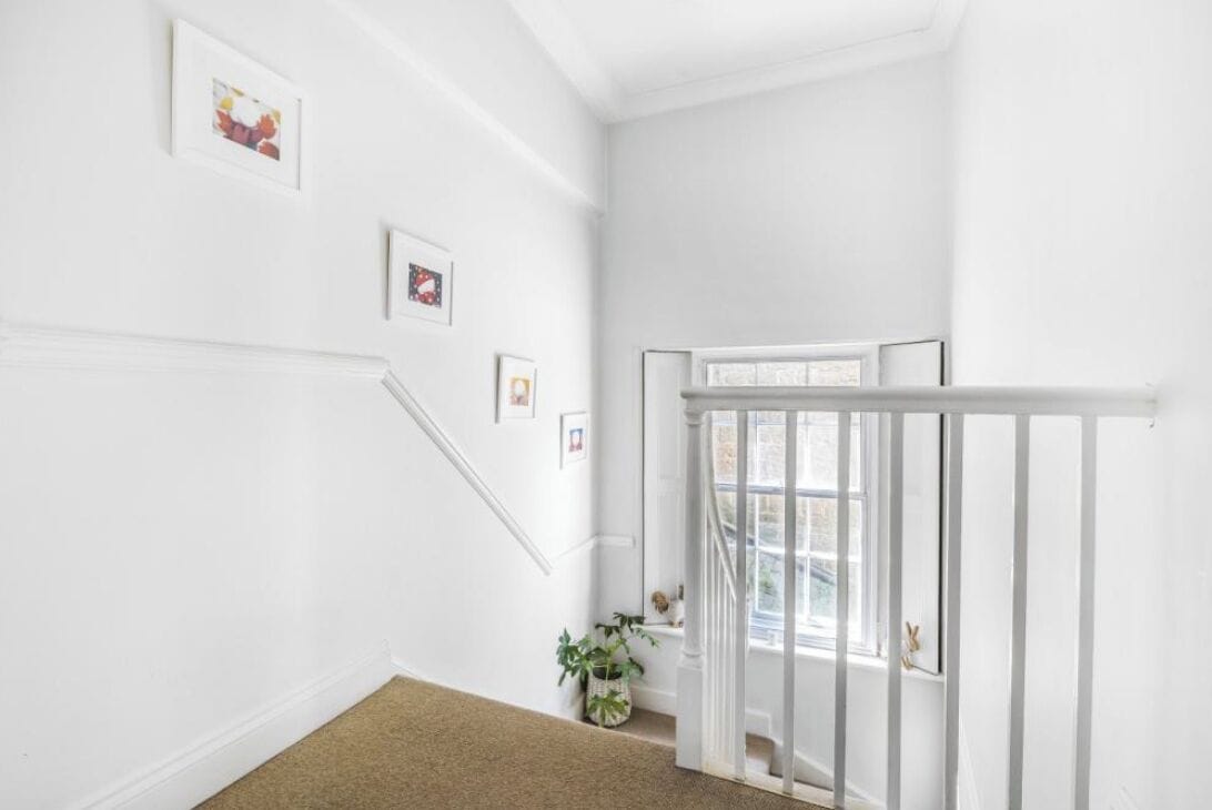 Un intérieur aux murs blancs, avec un escalier, des tableaux accrochés au mur et une plante en pot.