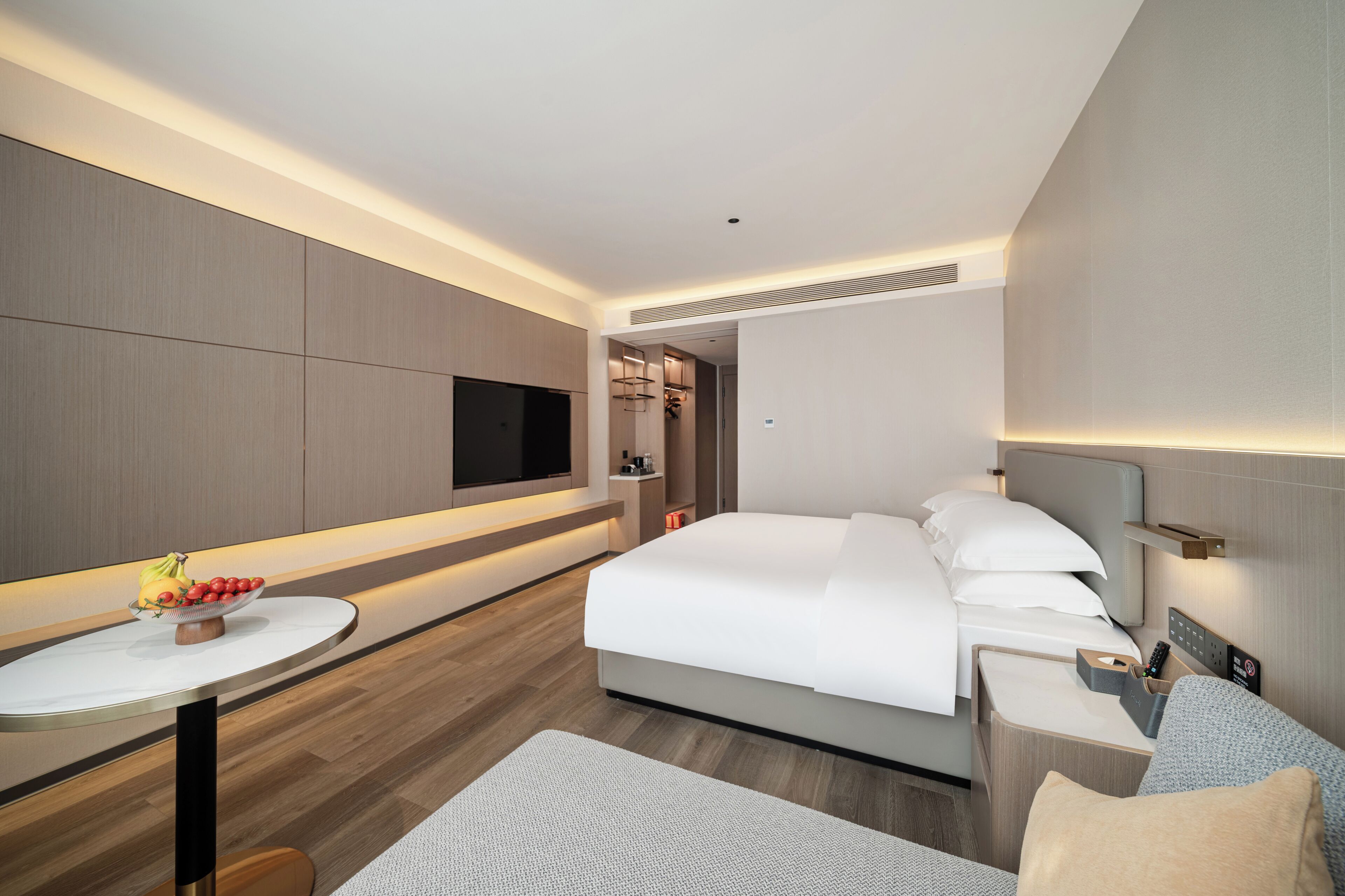 A modern hotel room with a bed, bedside table, TV, and a small seating area.