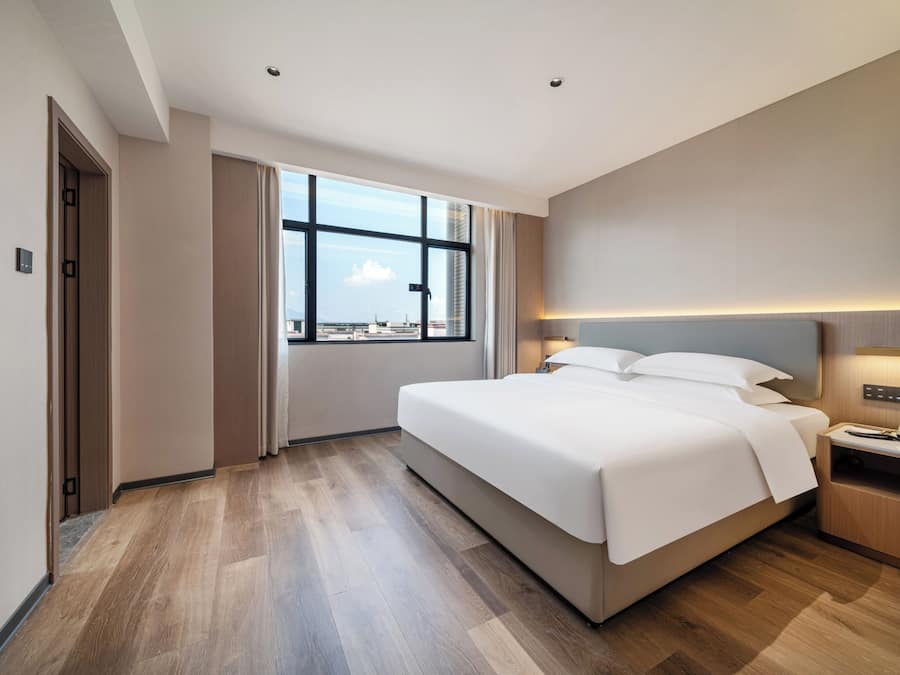 Quarto de hotel com uma cama grande, uma mesa com luminária, uma janela com vista e uma porta.