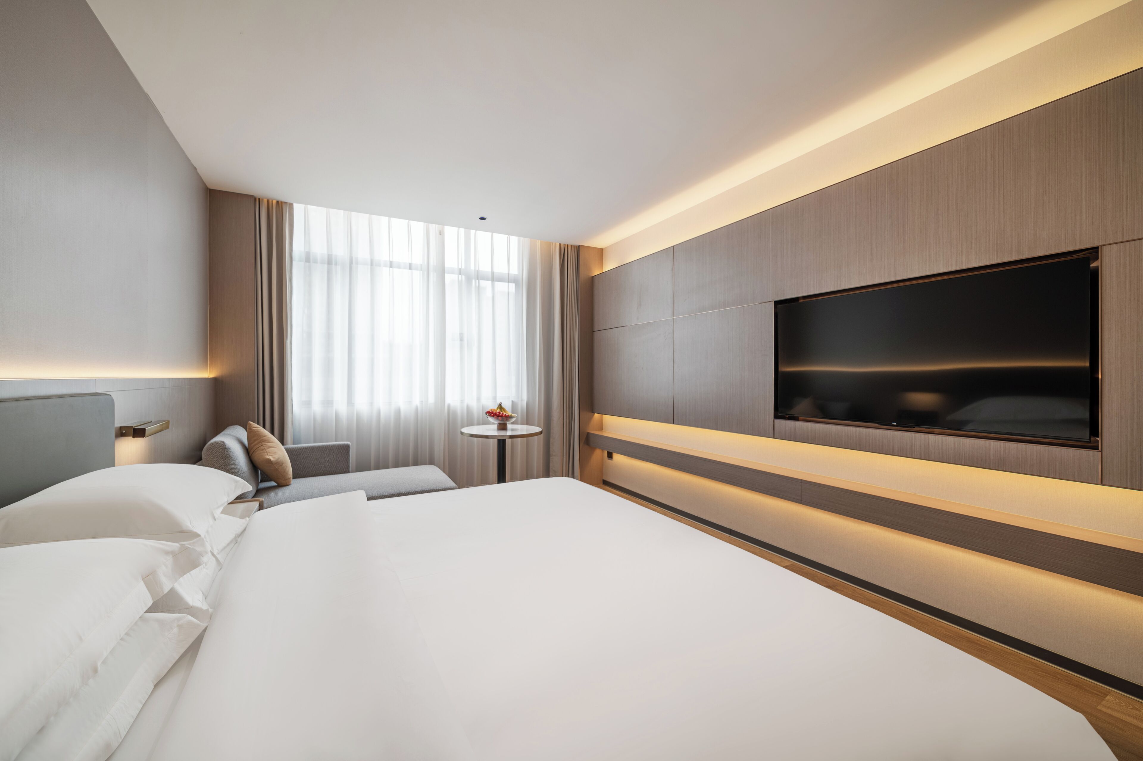 A modern hotel room with a large bed, a flat-screen TV mounted on the wall, and a small round table with a bowl of fruit.