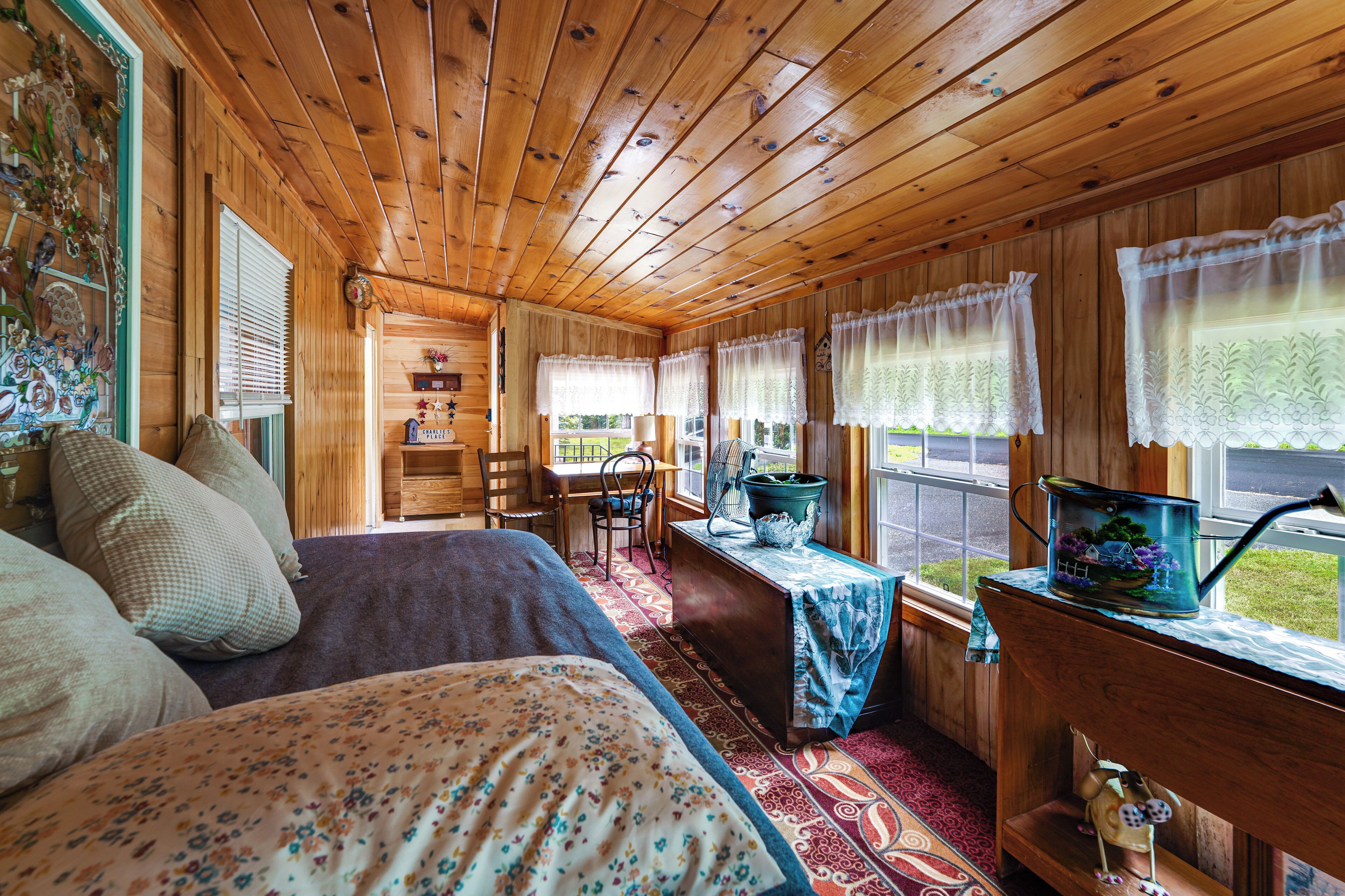 A wooden cabin interior with a bed, a table with a plant, and a window with curtains.