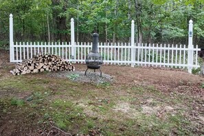 Ein Holzstapel, ein Metallgrill und ein weißer Zaun in einem bewaldeten Gebiet.