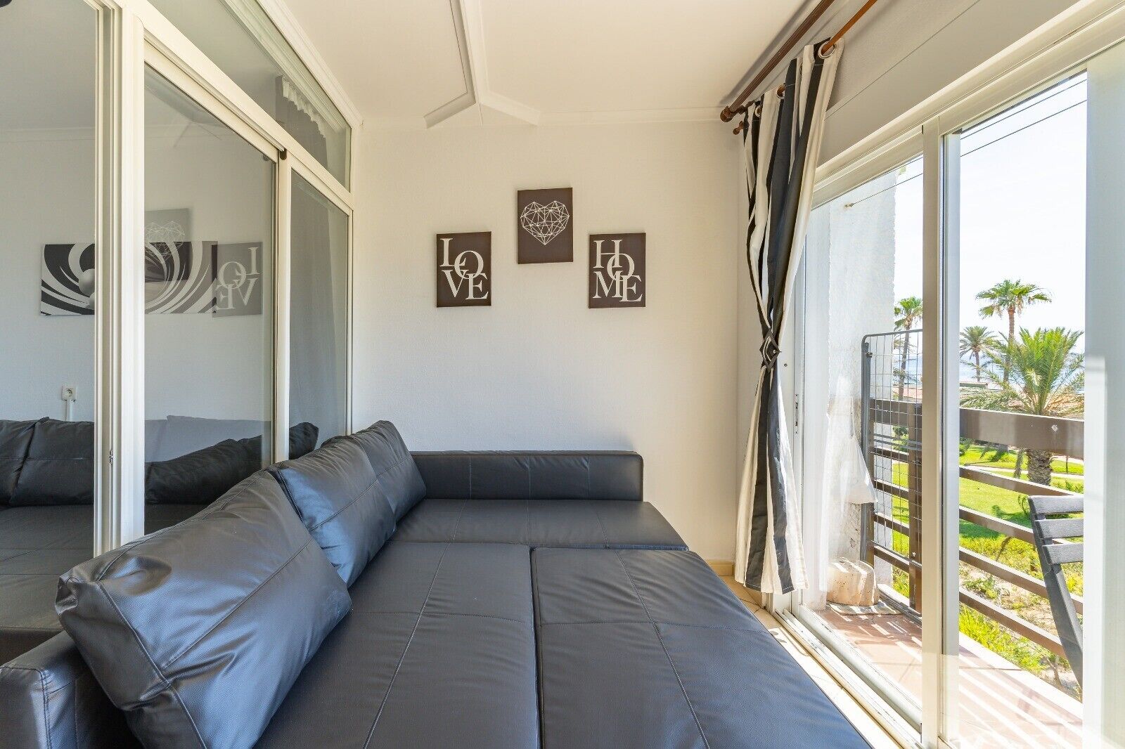 En moderne stue med en grå hjørnesofa, en glasdør der fører udendørs, og en balkon med udsigt til palmer.