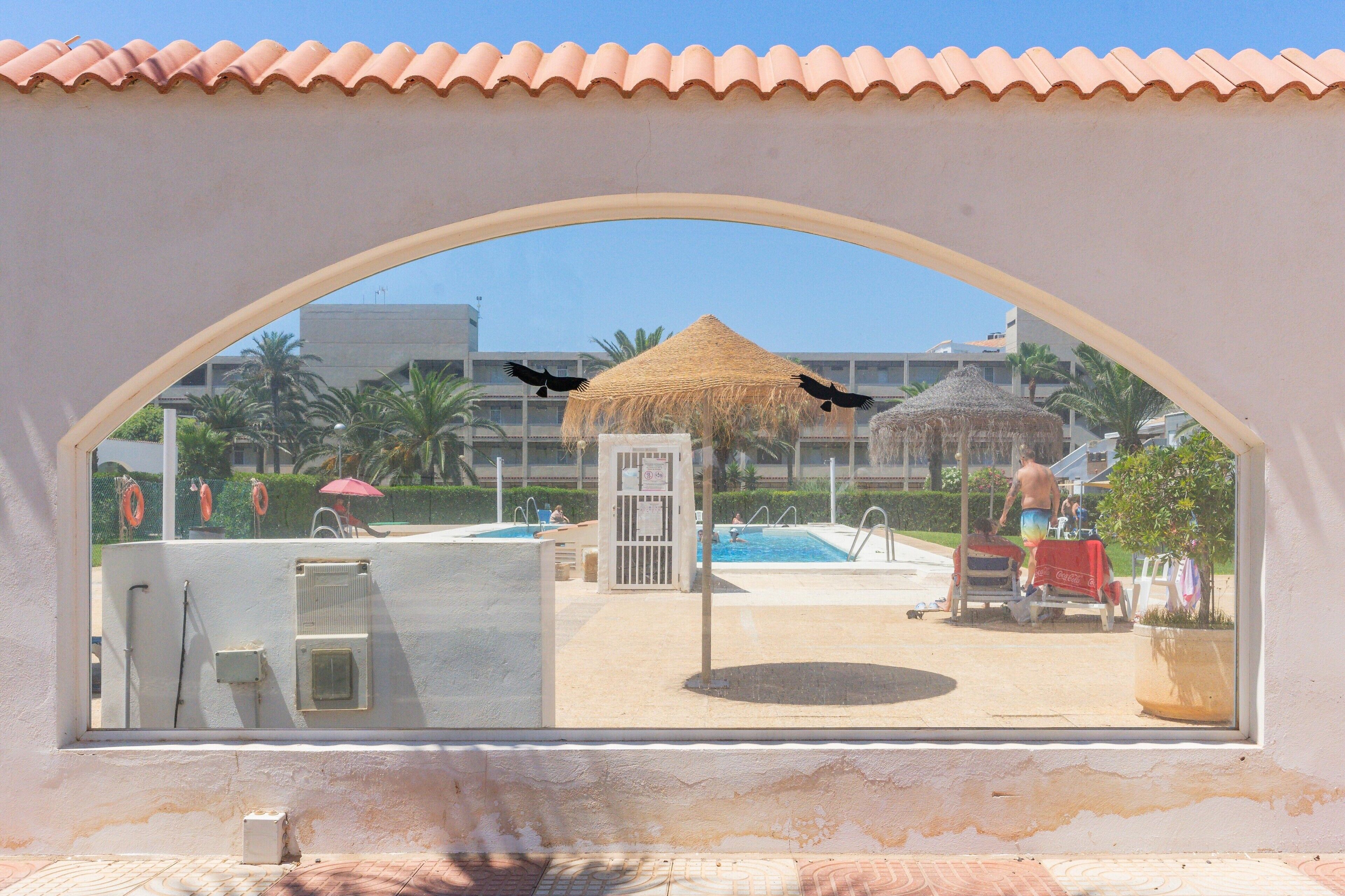Vista a través de un ventanal arqueado que muestra un área de piscina con una sombrilla de paja, un puesto de salvavidas y un edificio al fondo.