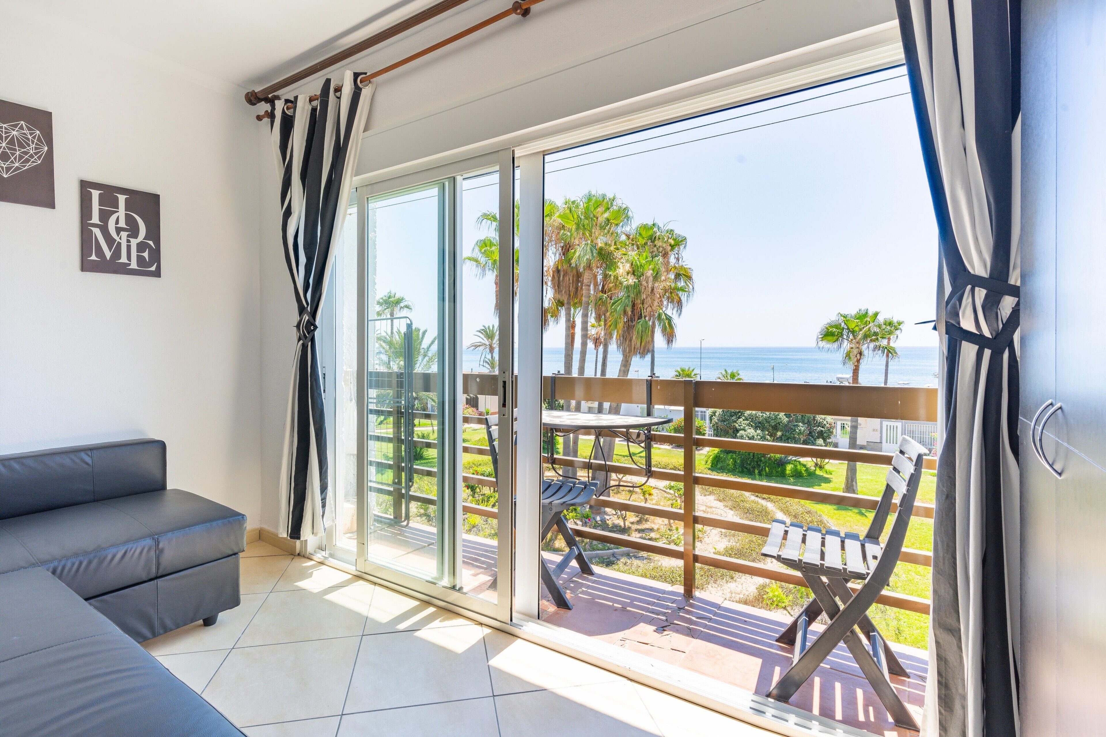 Una habitación moderna con un sofá gris, un amplio ventanal corredero que da a un balcón con vistas a palmeras y al mar.