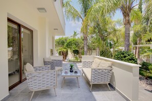 Une terrasse dotée de meubles en osier, d’une table avec des tasses et une plante, et offrant une vue sur des palmiers et une végétation luxuriante.
