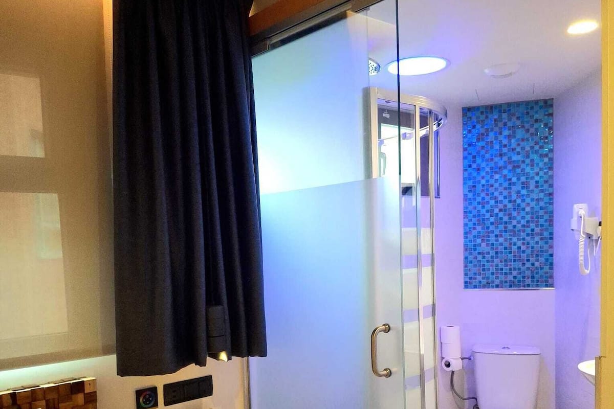Quarto de hotel com cama, mesa de cabeceira, chuveiro com box de vidro, sanita e parede com design de mosaico.
