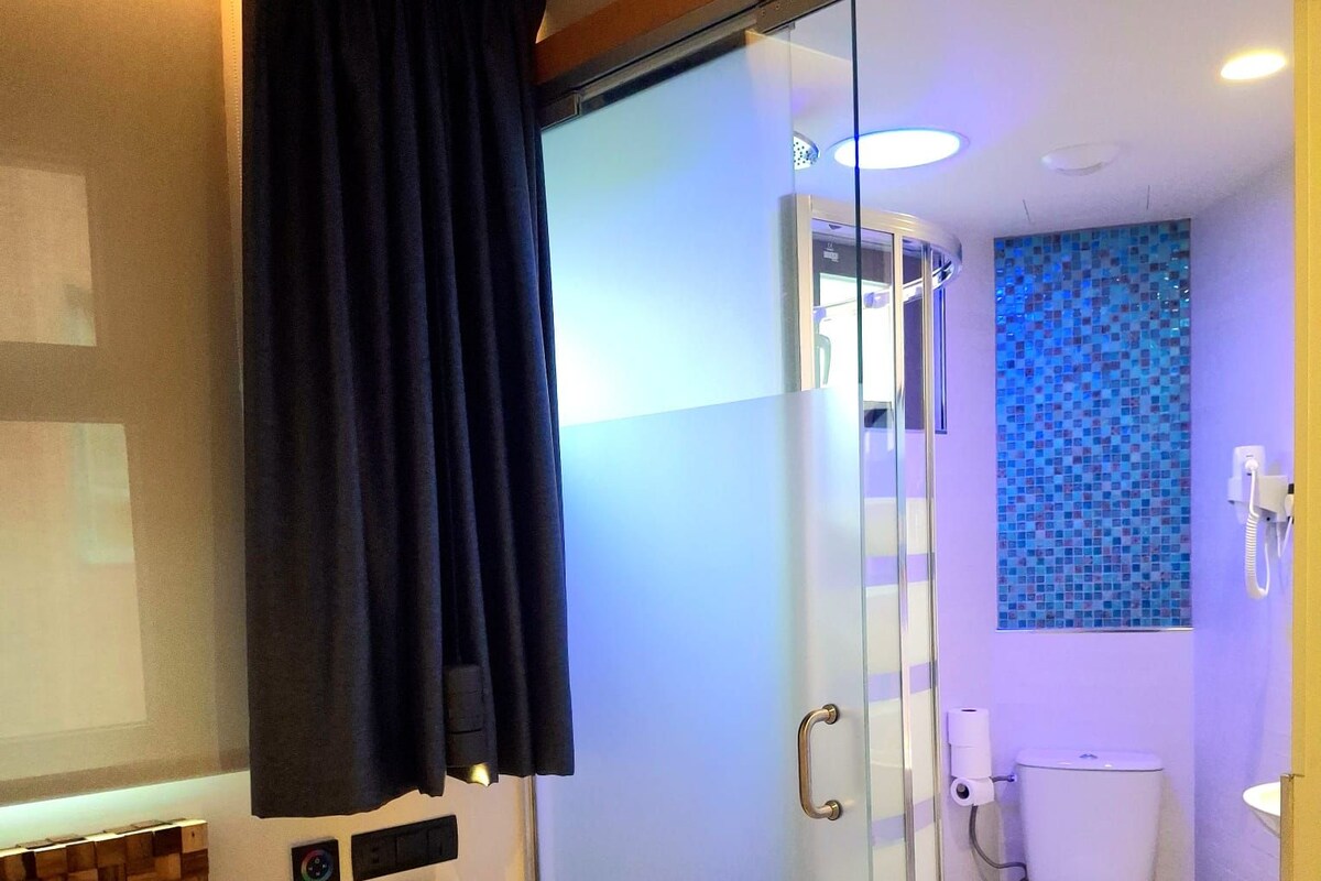 A hotel room with a bed, a bedside table, a glass-enclosed shower, a toilet, and a wall with a mosaic tile design.