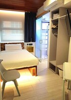 A hotel room with a bed, a chair, a desk, a wardrobe, and a sliding glass door leading to a balcony.