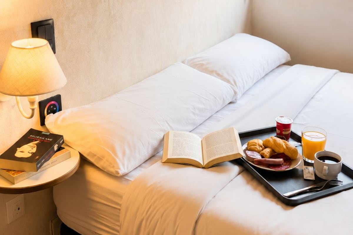 A neatly made bed with white linens, a bedside table with a lamp, an open book, a tray with croissants, juice, and coffee.
