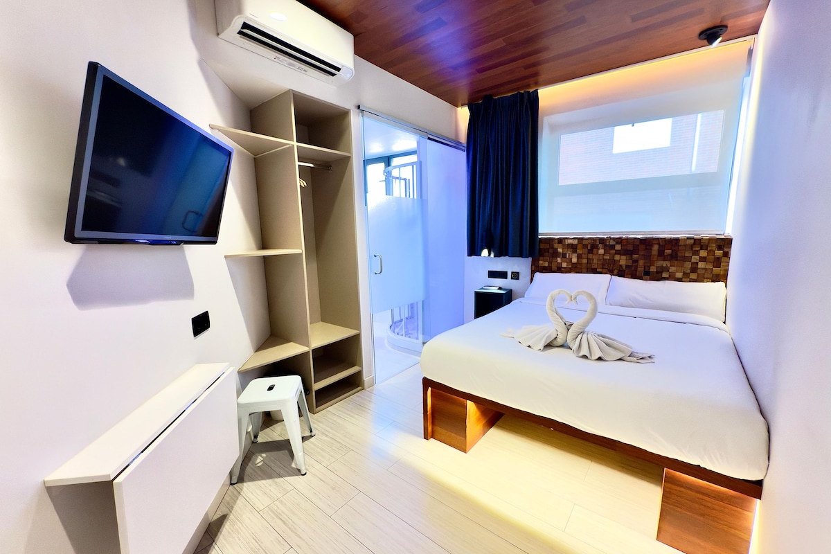 A modern hotel room with a bed, a TV, a wardrobe, and a small stool.