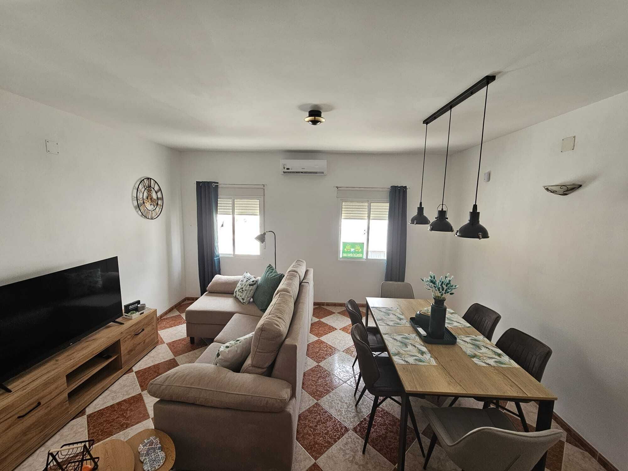 Een woonkamer met een bank, televisie, eettafel, stoelen en een plafondventilator.