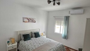 Un dormitorio con cama, mesita de noche, aire acondicionado mural, ventilador de techo y ventana con cortinas.