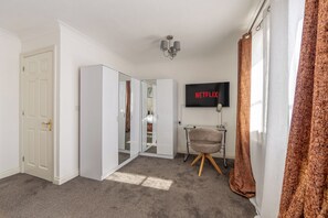 A hotel room with a wardrobe, a television displaying the Netflix logo, a chair, and a curtain. - Luxury 4 Bedroom Home, Sleeps 10, Garden, Free Parking, Hornchurch, Near London (Hornchurch)