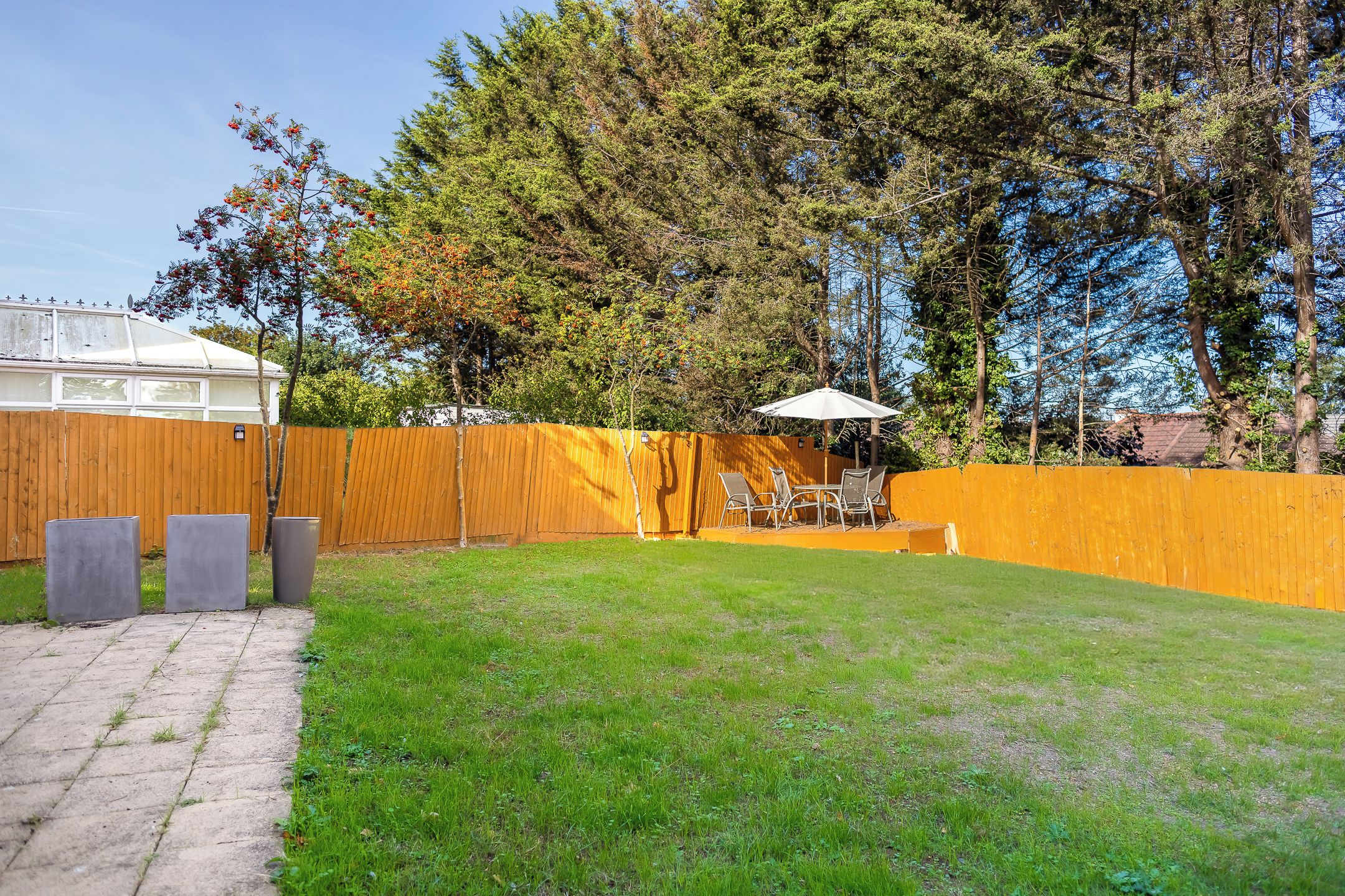 Un patio trasero cercado con césped, un patio con sillas y una mesa, y una valla de madera.