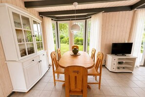 A dining area with a wooden table and chairs, a cabinet with glass doors, a television on a stand, and a view of greenery outside. - Skandinavisches Ferienhaus mit Privatsauna im Hafendorf Müritz (Granzow)