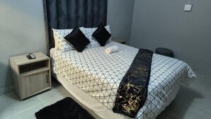 A bedroom with a bed, a bedside table, a nightstand, a black rug, and a wall-mounted artwork. - Acama Guest House 455 Long rd, Welkom, Free State (Welkom)
