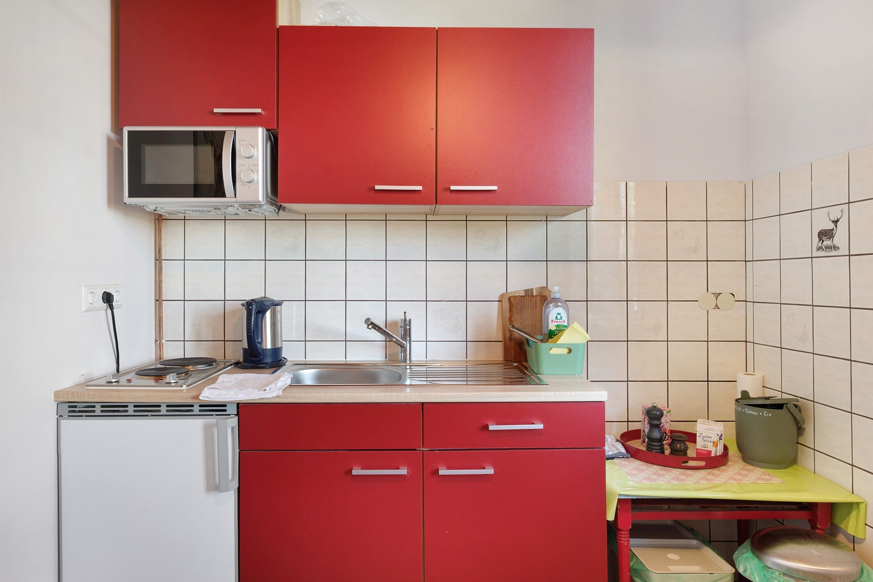 A compact kitchen with red cabinets, a microwave, a sink, and a small table with a kettle and a bottle of dish soap.