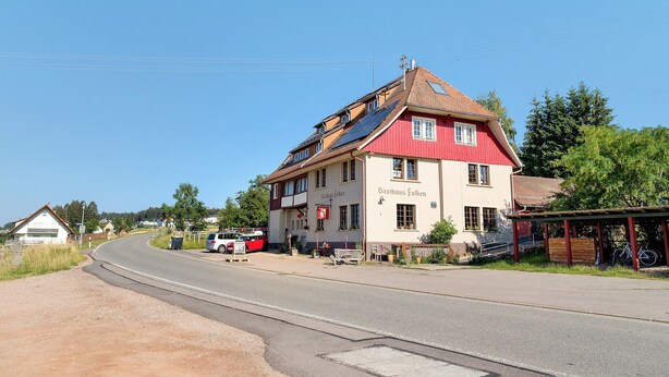 A red and white building with solar panels on the roof, a sign reading 'Gasthaus Falken,' and a bicycle parked outside. - Tiny Home “Bauwagen auf dem Falkenhof” – Nature Retreat with Shared Kitchen (Eisenbach)