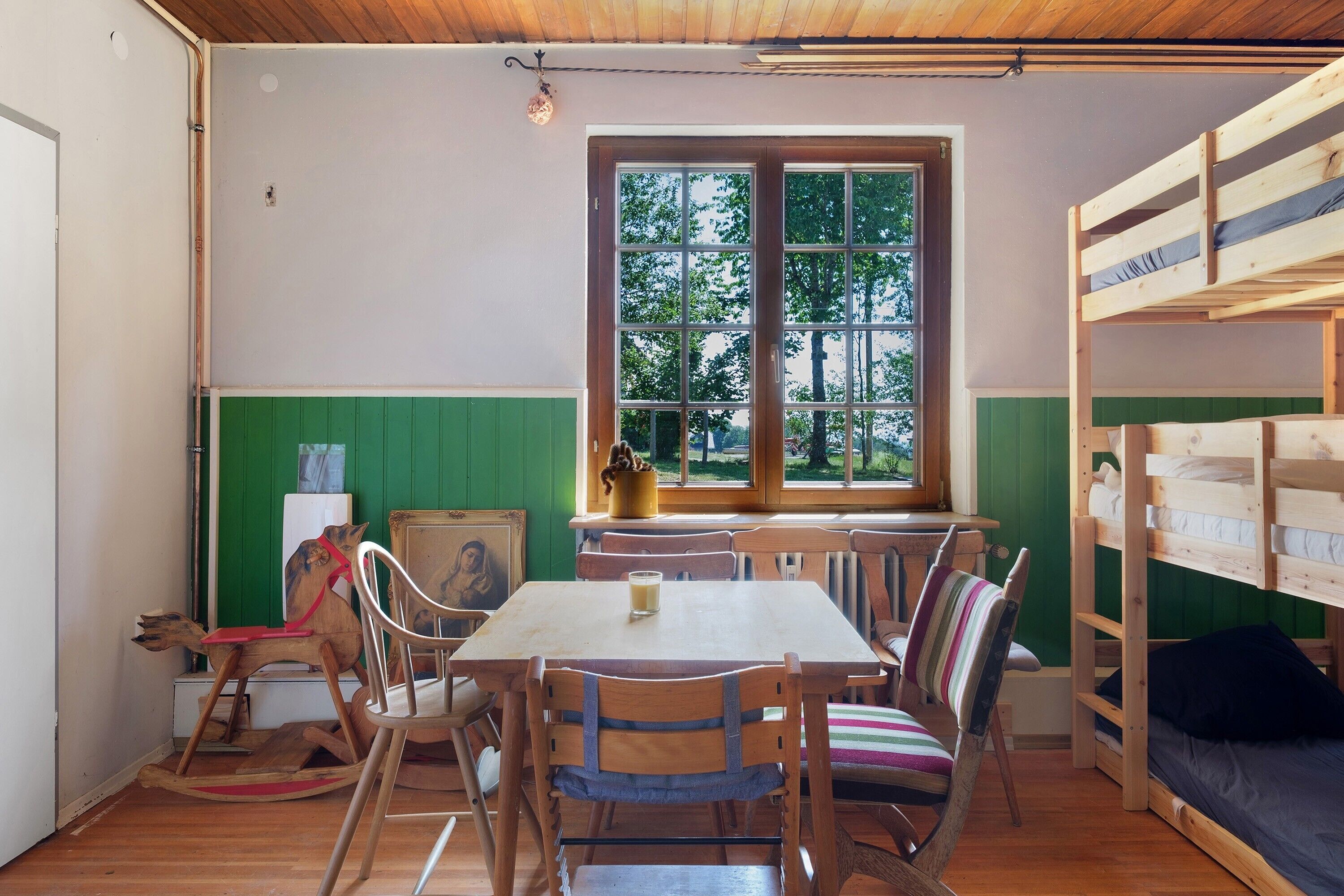 Ein Zimmer mit Holztisch, Stühlen, einem Etagenbett und einem Fenster mit Blick auf Bäume.