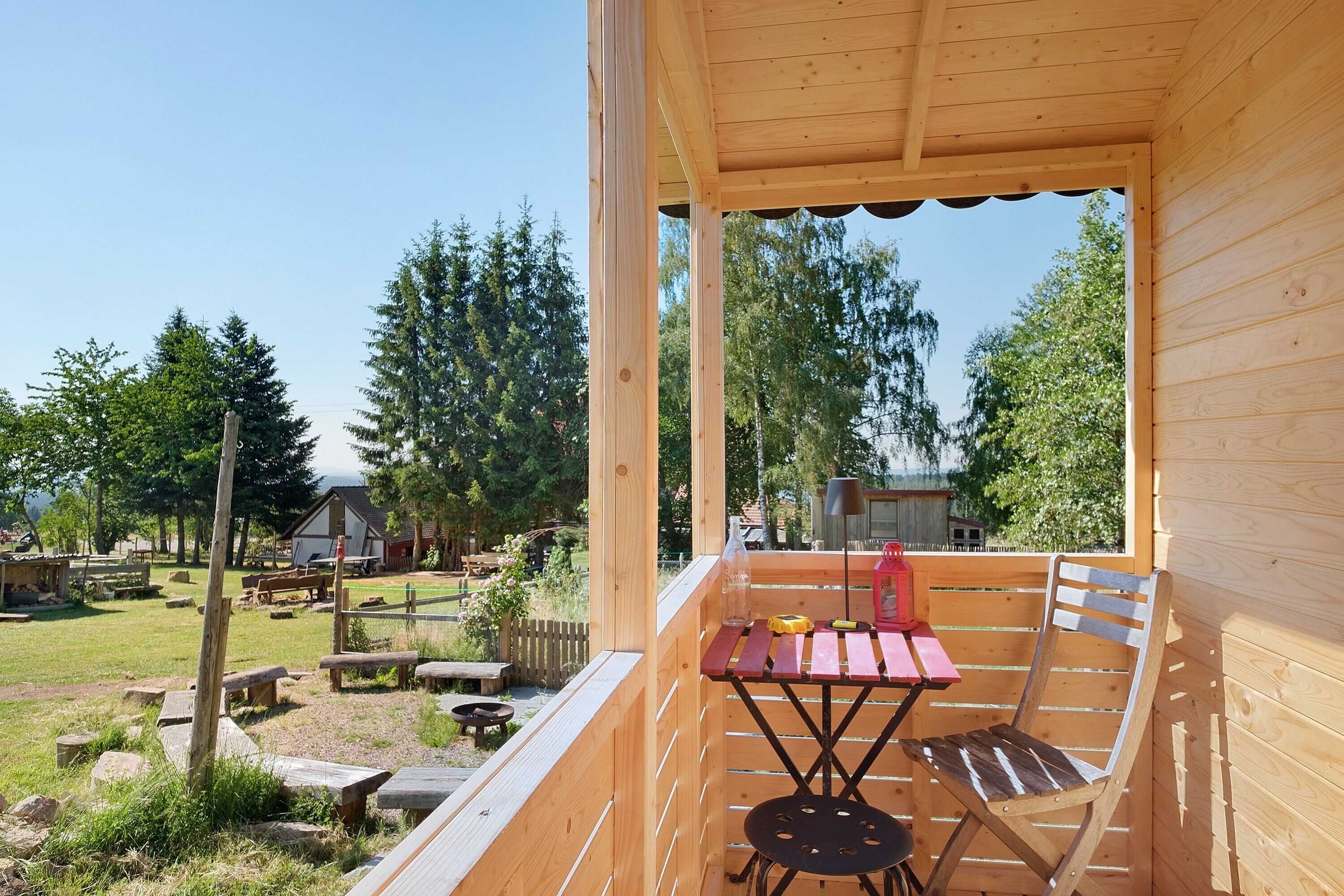 Eine Holzdielen-Terrasse mit Tisch und Stühlen, mit Blick auf eine Rasenfläche mit Bäumen und einem Gebäude im Hintergrund.
