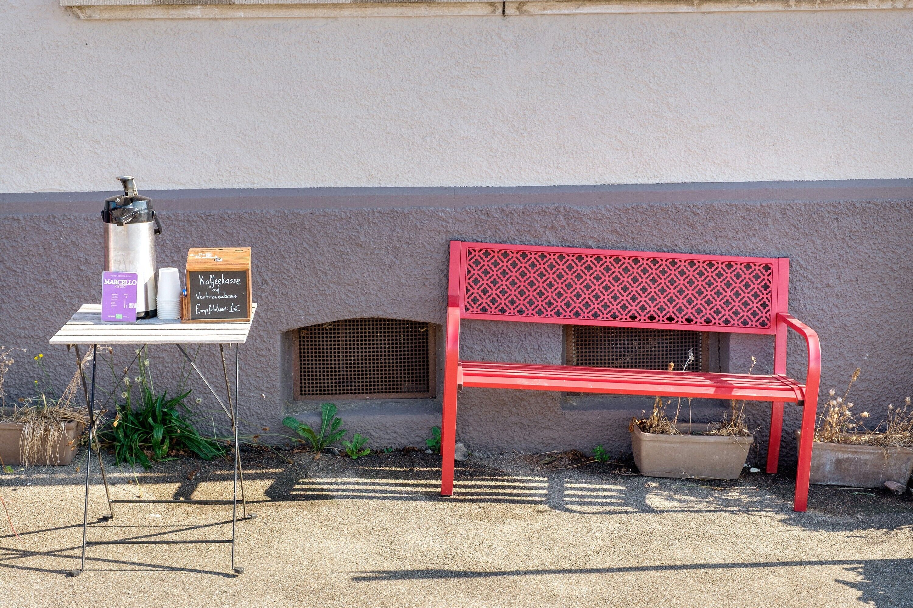 Eine rote Bank, eine Kaffeemaschine und ein Schild an einer Hauswand.