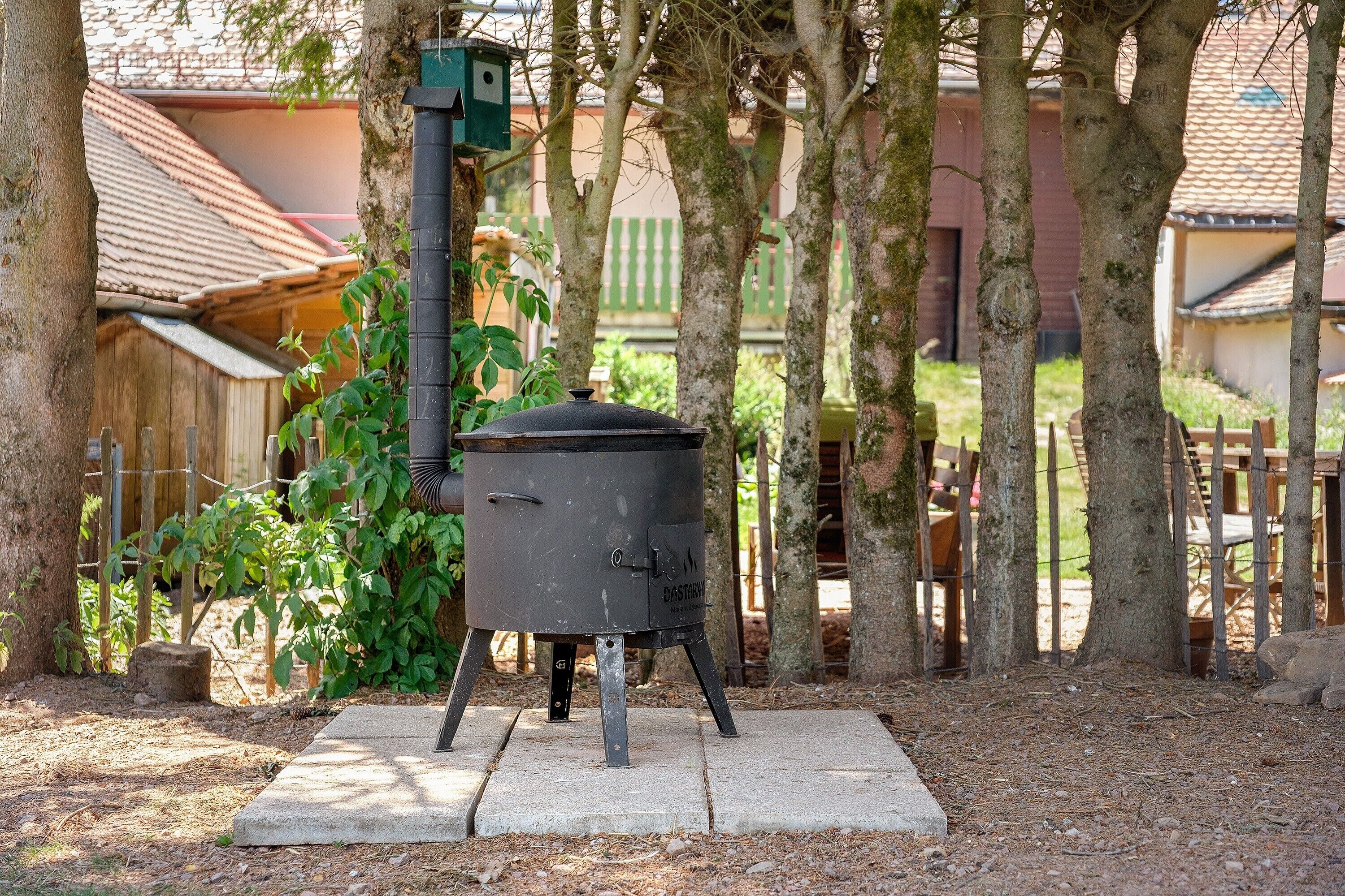 Una estufa de cocina portátil para exteriores con chimenea, ubicada sobre una base de concreto rodeada de árboles.