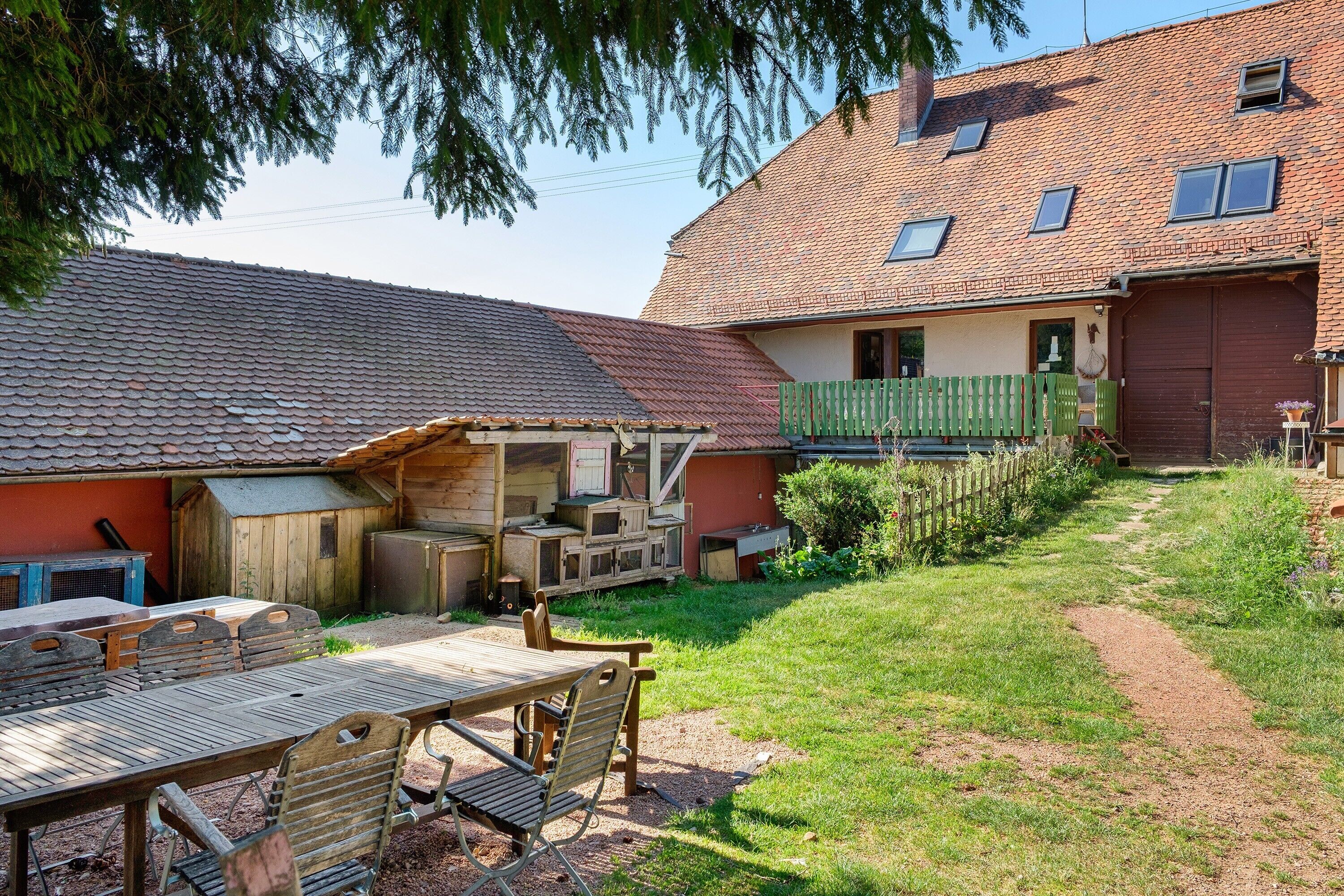 Un espace extérieur rustique avec une table et des chaises en bois, un coin salon abrité et un jardin clôturé.