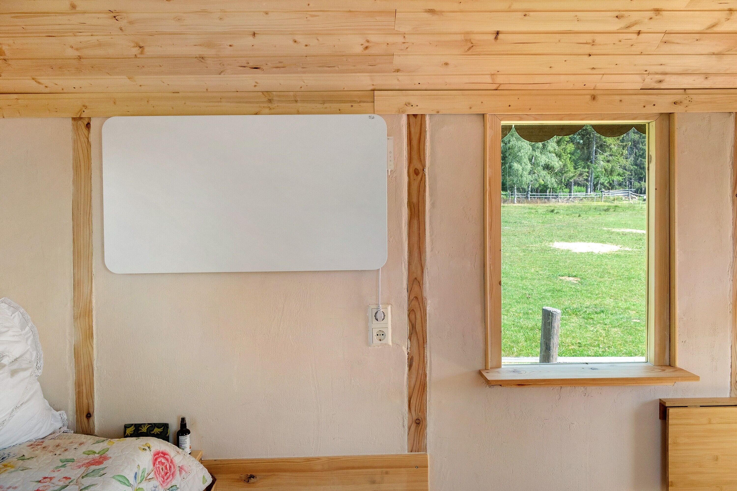 A bedroom with a wooden ceiling, a bed with a floral bedspread, a window with a view of a grassy area, and a whiteboard on the wall.